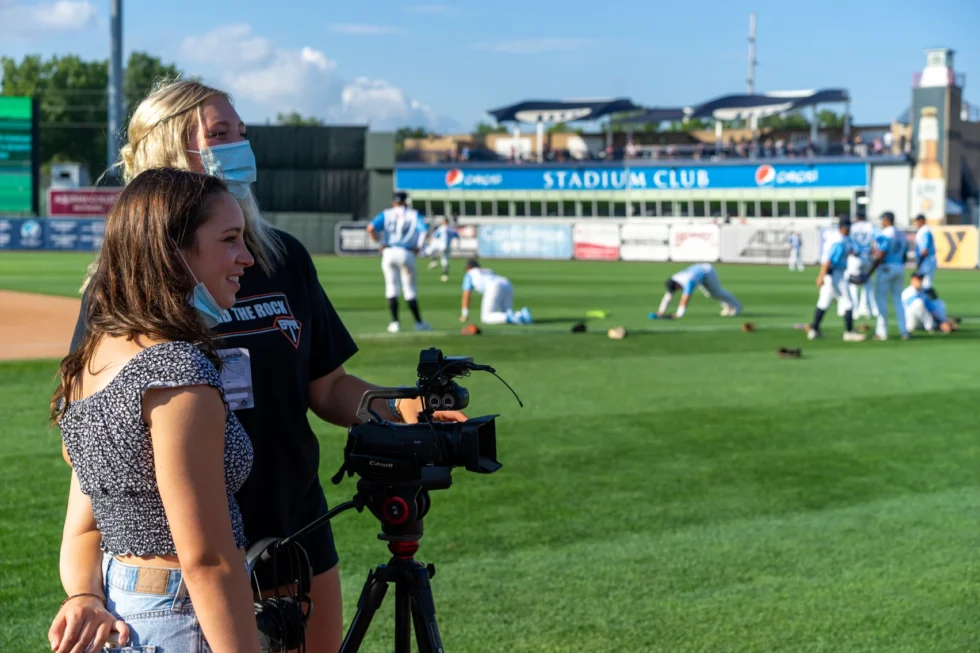 Student operating a camera on a tripod at the ballpark